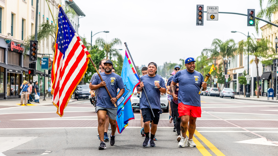 Men run together on a palm tree-lined street; one man carries a torch