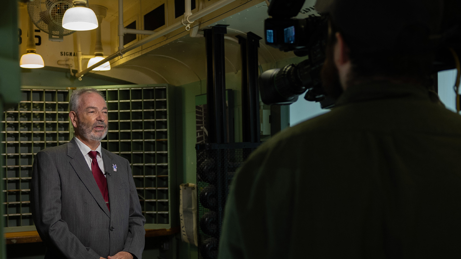 A man stands in a vintage rail car and speaks to another man holding a TV camera