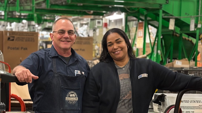 A man and a woman stand among mail transport equipment on the workroom floor of a postal processing plant