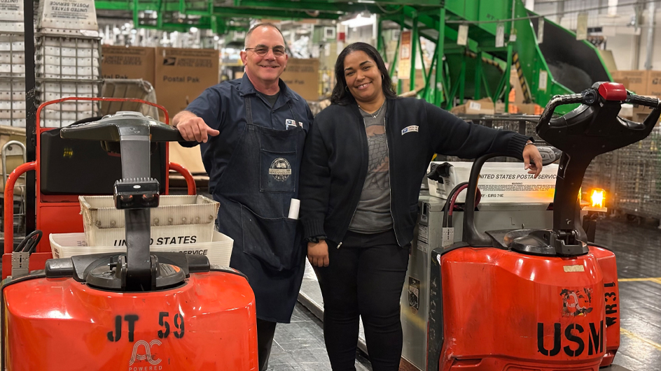 A man and a woman stand among mail transport equipment on the workroom floor of a postal processing plant