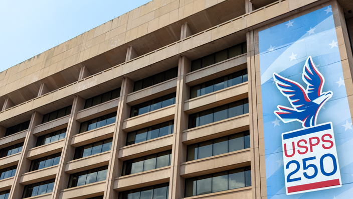 A large banner hanging on the side of an office building. The banner shows an illustration of an eagle about to take wing and the words “USPS 250.”
