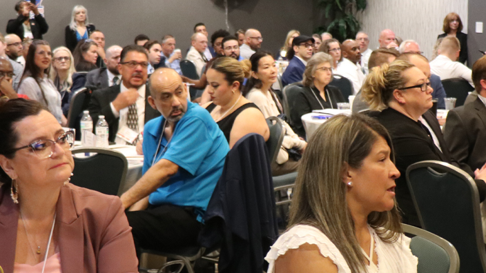 People sit at tables in a hotel ballroom and listen to an unseen speaker