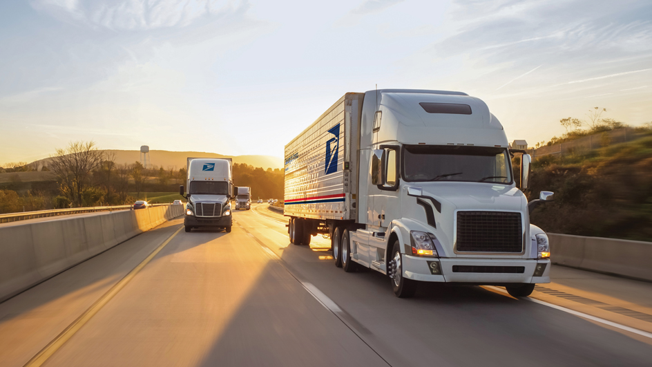 Two USPS branded tractor-trailer trucks move along a highway.