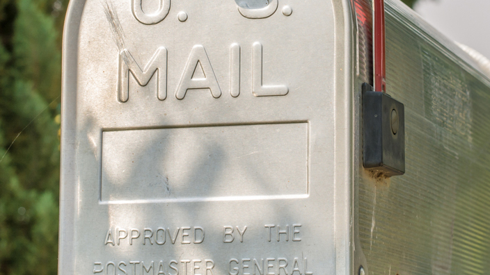 A metal mailbox on a suburban street, bearing the words “U.S. Mail” and “Approved by the Postmaster General.”