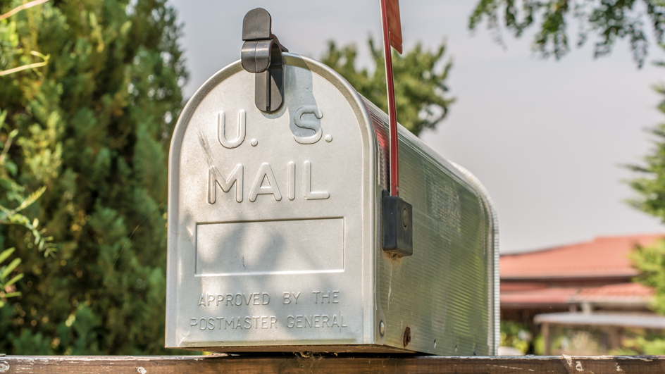 A metal mailbox on a suburban street, bearing the words “U.S. Mail” and “Approved by the Postmaster General.”