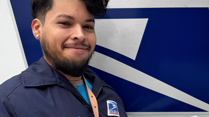 A smiling man wearing a postal uniform stands next to a mail delivery truck