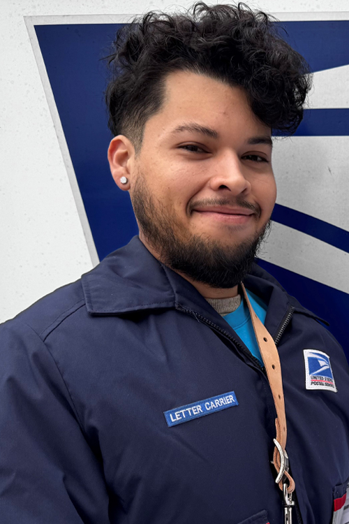 A smiling man wearing a postal uniform stands next to a mail delivery truck