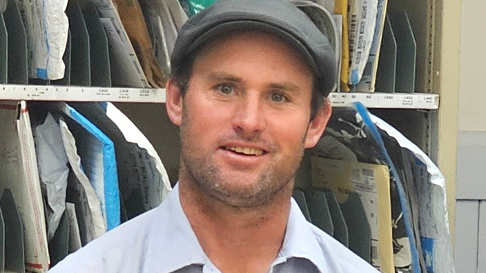 A man wearing a USPS uniform stands in a Post Office workroom and smiles