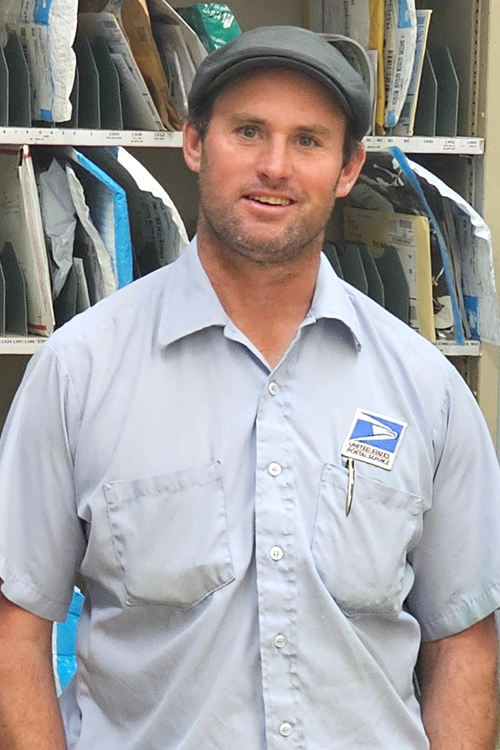 A man wearing a USPS uniform stands in a Post Office workroom and smiles