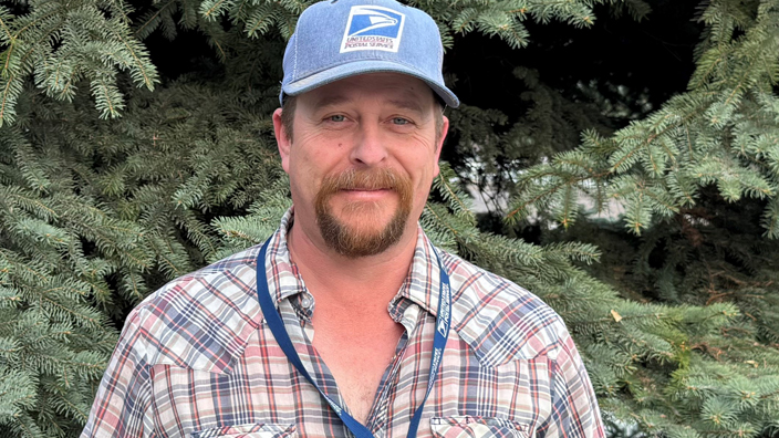 A man wearing a USPS baseball cap and a plaid shirt stands in front of a large tree