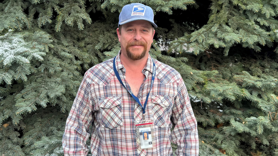 A man wearing a USPS baseball cap and a plaid shirt stands in front of a large tree