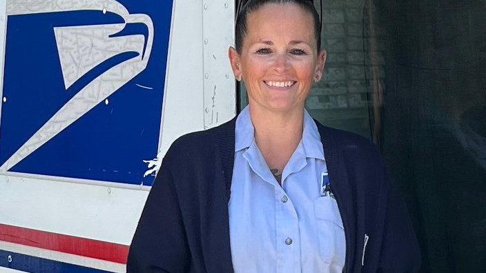 A woman in a postal uniform smiles while standing next to a mail delivery truck