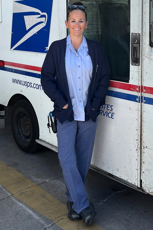 A woman in a postal uniform smiles while standing next to a mail delivery truck