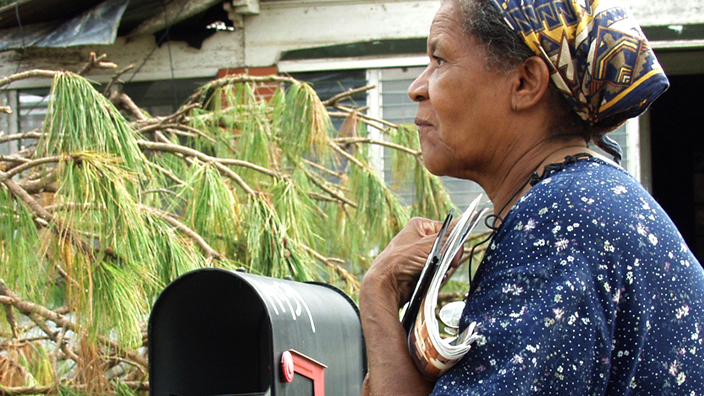 A woman stands in the front yard of a ruined home and holds a stack of mail close to her body