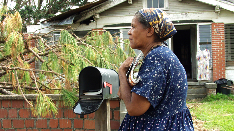 A woman stands in the front yard of a ruined home and holds a stack of mail close to her body