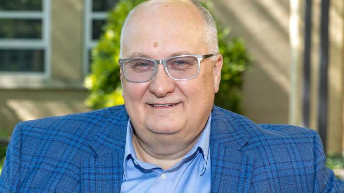 A man wearing a sportcoat smiles as he sits on a bench in a park-like setting