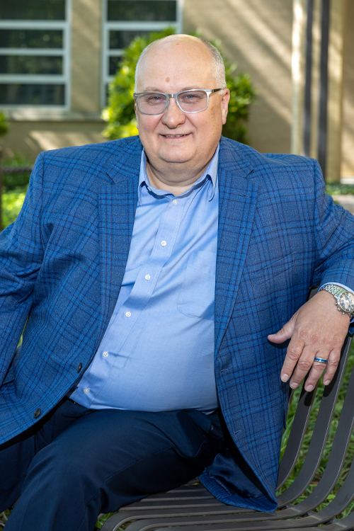 A man wearing a sportcoat smiles as he sits on a bench in a park-like setting
