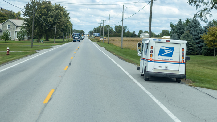 A mail delivery truck stops at a mailbox along a rural road