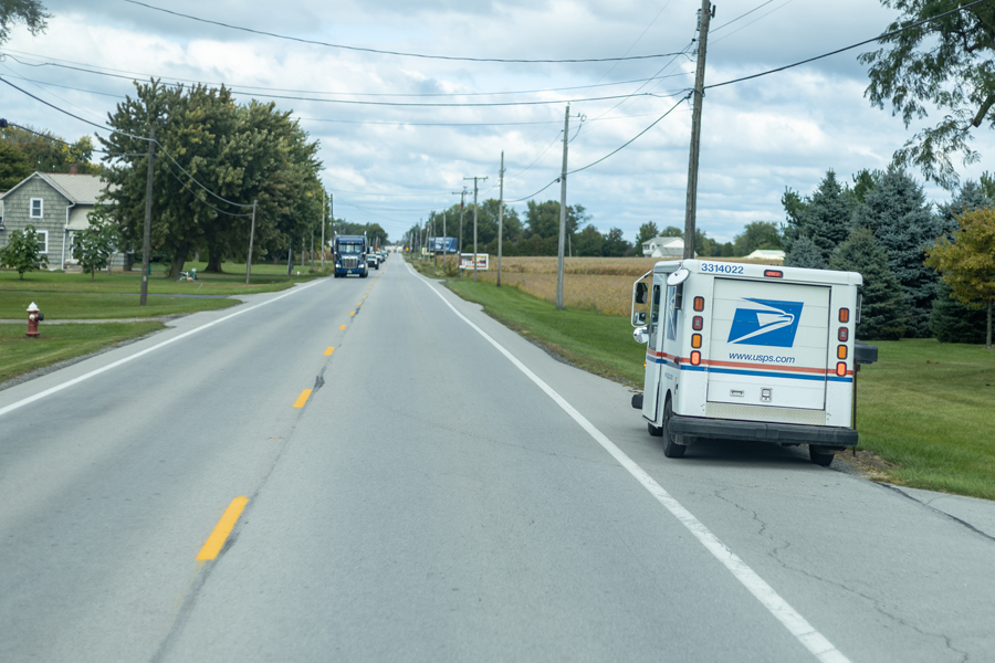 A mail delivery truck stops at a mailbox along a rural road