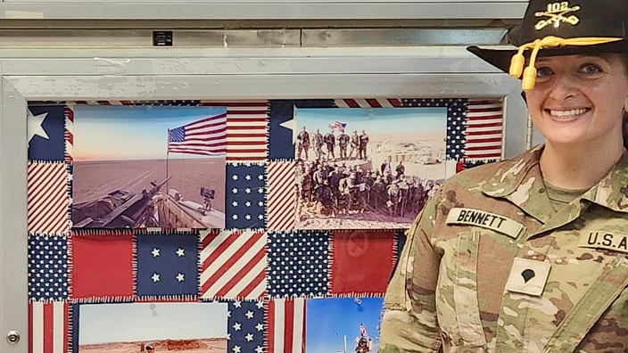 A woman in Army fatigues stands next to a flag in a glass display case