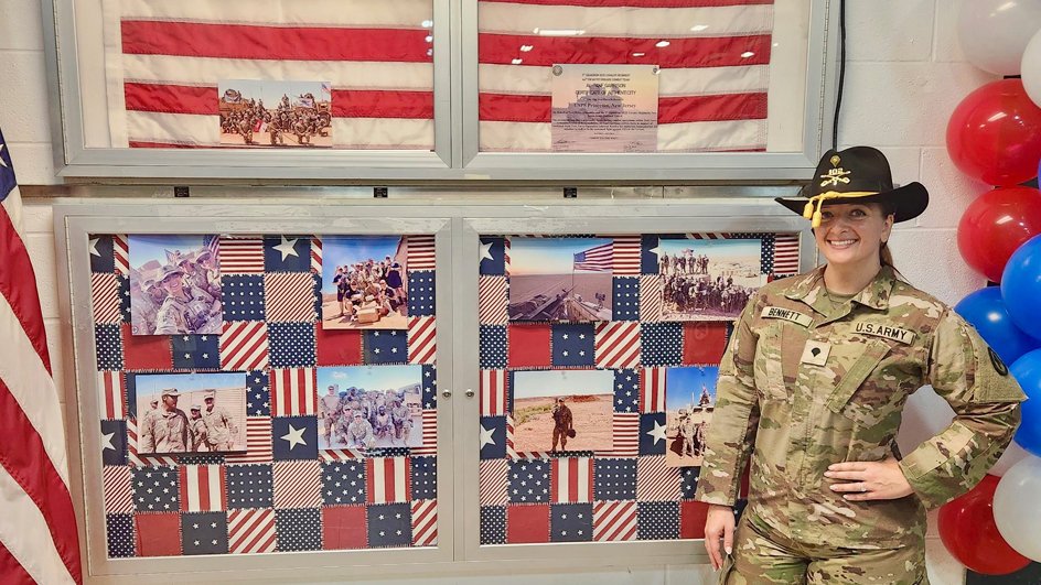 A woman in Army fatigues stands next to a flag in a glass display case