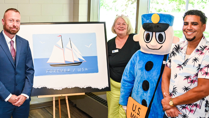 A group of people surround an enlarged image of a stamped postcard on an easel.