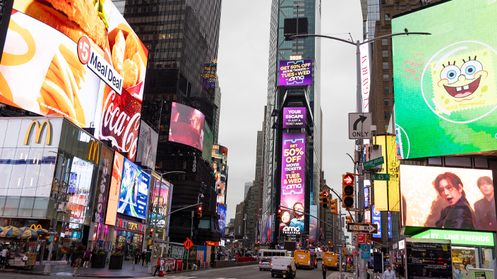 Pedestrians cross a New York City street while videos play on screens above