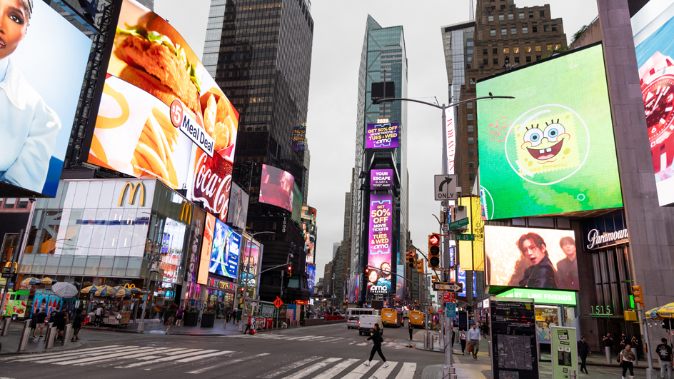 Pedestrians cross a New York City street while videos play on screens above