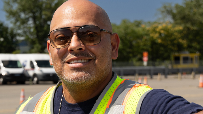 A man wearing a USPS-branded reflective safety vest stands on pavement with Postal Service delivery vehicles looming in the distance