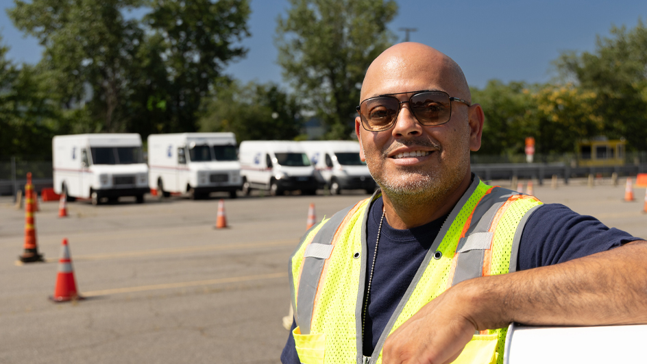 A man wearing a USPS-branded reflective safety vest stands on pavement with Postal Service delivery vehicles looming in the distance