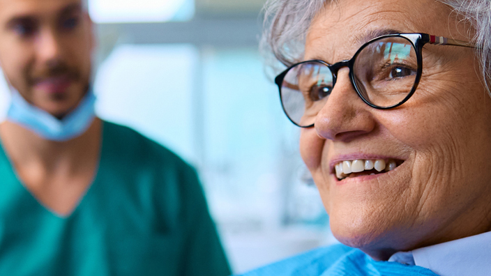 A dentist watches as a woman in his examining chair holds a mirror to look at her smile