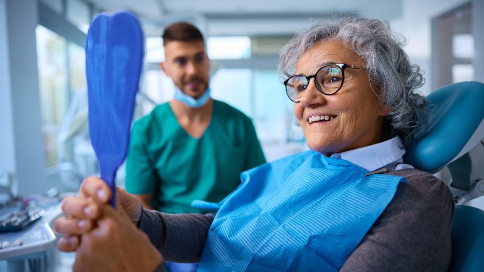 A dentist watches as a woman in his examining chair holds a mirror to look at her smile