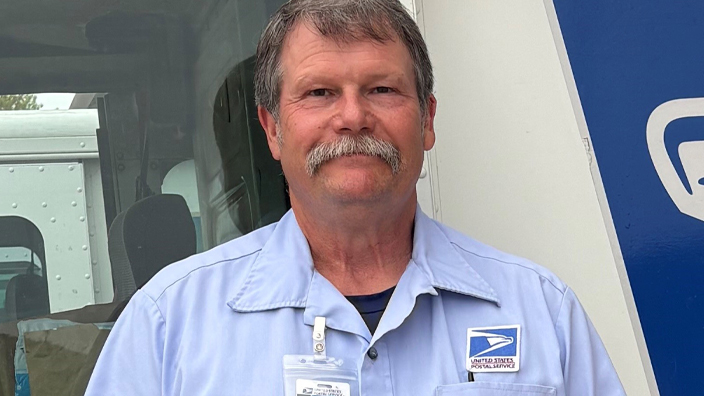 A man wearing a Postal Service uniform stands next to a delivery truck