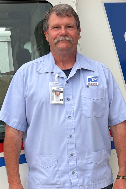 A man wearing a Postal Service uniform stands next to a delivery truck