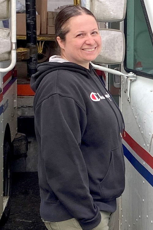 A woman wearing a hoodie smiles as she stands next to a Postal Service delivery truck