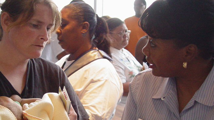 A woman holding a baby speaks to a uniform-clad postal worker under a large tent
