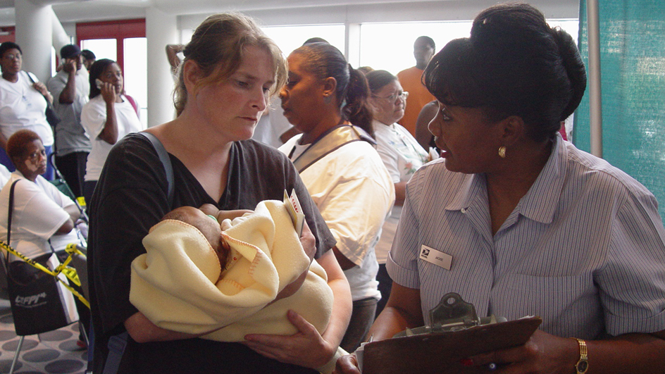 A woman holding a baby speaks to a uniform-clad postal worker under a large tent