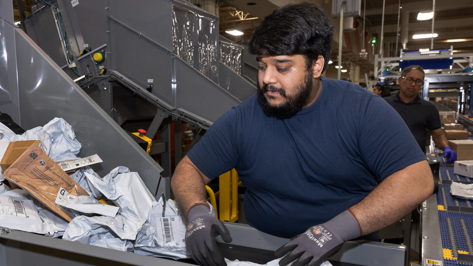 A male USPS employee handles mail in a sorting and distribution facility.