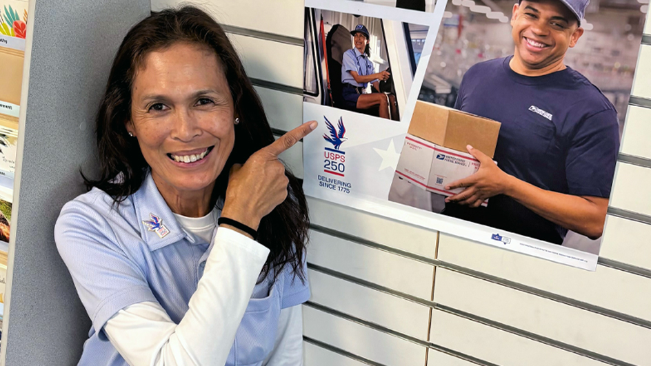 A woman wearing a postal uniform smiles and points to a poster hanging in a Post Office lobby.
