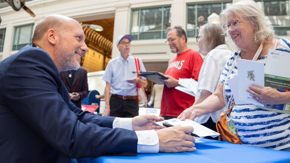 A smiling man sits at a table and signs booklets