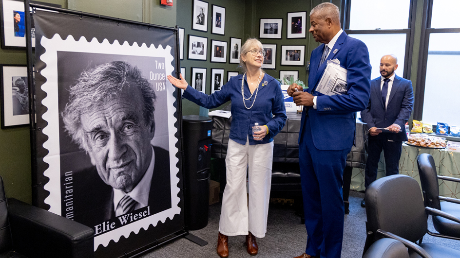 A woman stands near a poster displaying an image of Elie Wiesel and speaks to a man in a business suit