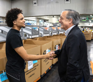 A man in a sport coat shakes hands with a younger man dressed in a T-shirt and jeans