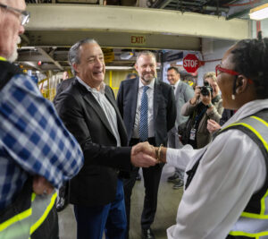 A man in a sport coat greets a woman wearing a reflective safety vest on the workroom floor of a postal processing plant.