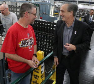 A man in a sport coat speaks with a man in a T-shirt on the workroom floor of a postal processing plant