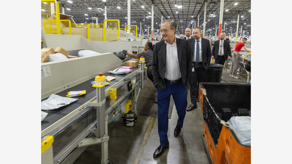 A man in a sport coat walks through a postal processing plant accompanied by men and women in business suits.