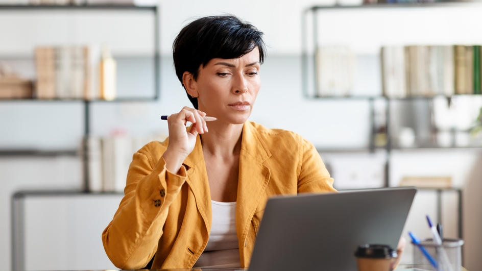 A woman with a contemplative expression sits at a desk and looks at a computer monitor
