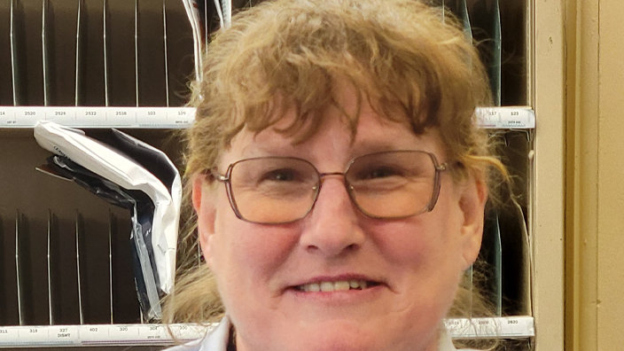 A woman wearing a Postal Service uniform stands and smiles in a Post Office workroom