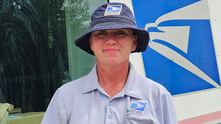 A smiling woman wearing a USPS hat stands next to a Postal Service delivery vehicle