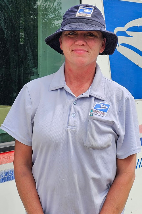 A smiling woman wearing a USPS hat stands next to a Postal Service delivery vehicle