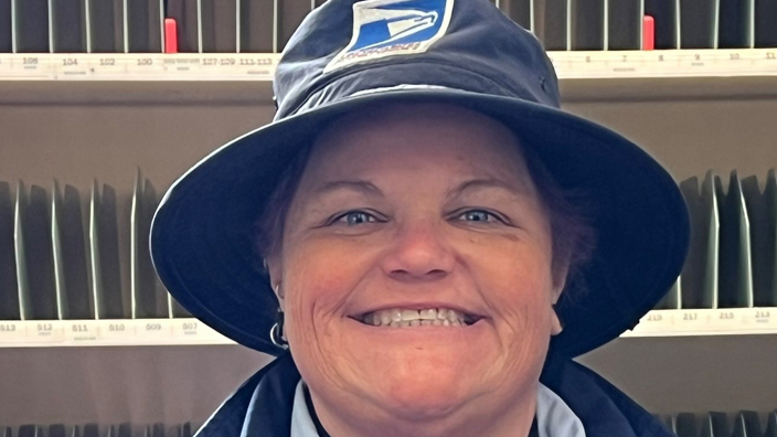 A woman wearing a Postal Service uniform stands and smiles inside a Post Office workroom